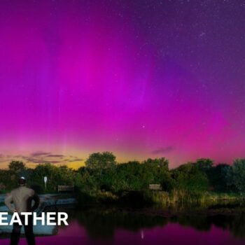 at night, in the foreground is a a man looking out across a body of water with trees on the horizon.  The night sky above is filled with different shades of purple of the northern lights.  A small streak of white light is seen in the top corner - a meteor.