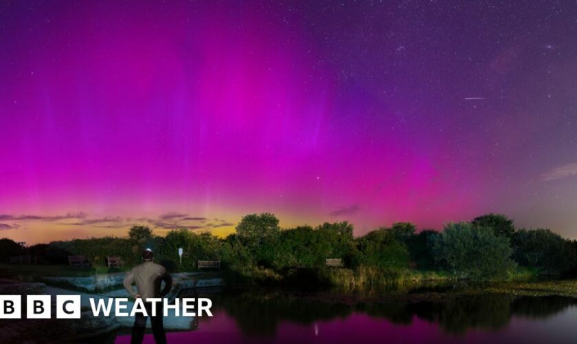 at night, in the foreground is a a man looking out across a body of water with trees on the horizon.  The night sky above is filled with different shades of purple of the northern lights.  A small streak of white light is seen in the top corner - a meteor.