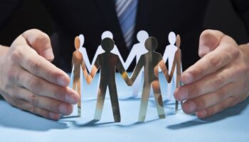 Businessman's Hands Protecting Team Of Paper People On Desk