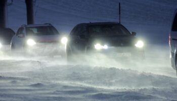 Autos mit eingeschalteten Scheinwerfern fahren in Kolonne über eine Straße mit Schnee und Glätte