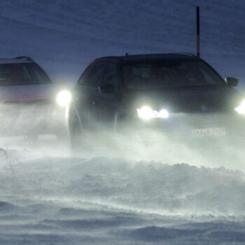 Autos mit eingeschalteten Scheinwerfern fahren in Kolonne über eine Straße mit Schnee und Glätte