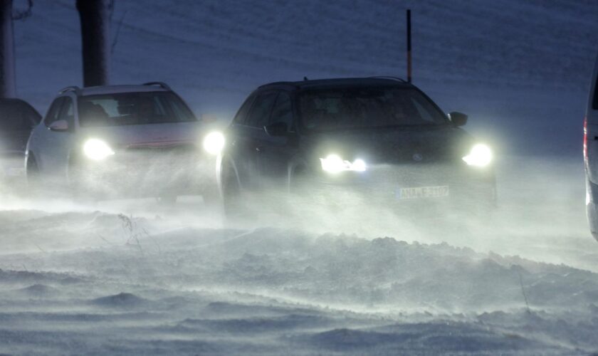 Autos mit eingeschalteten Scheinwerfern fahren in Kolonne über eine Straße mit Schnee und Glätte